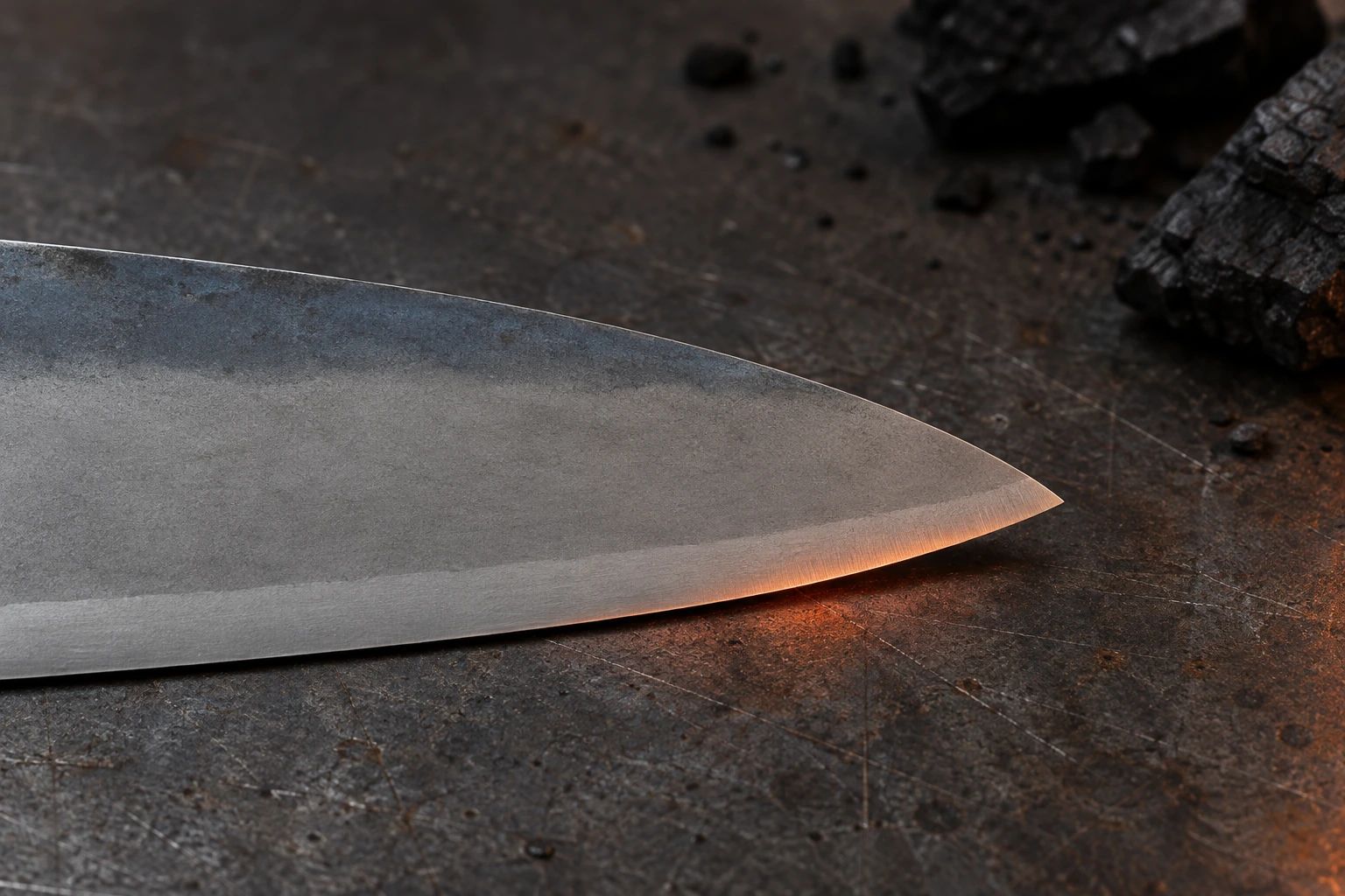 Close-up of the tip of a carbon-steel gyuto with a hazy kasumi finish on a steel workbench beside charcoal