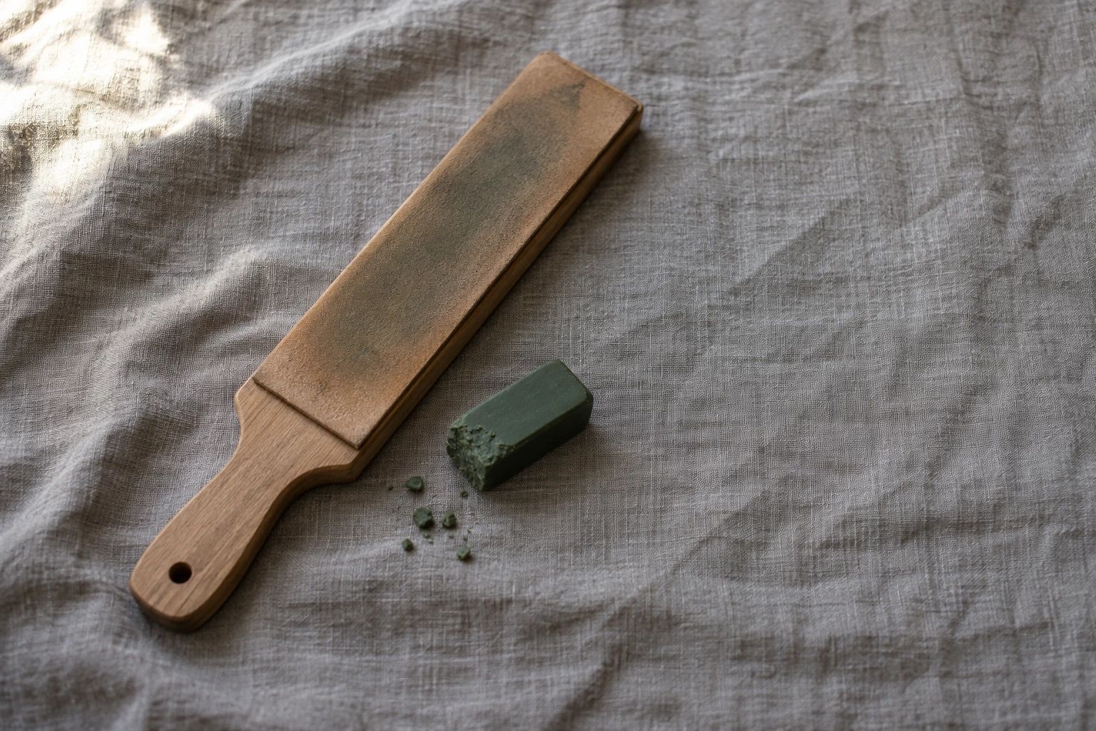 A leather strop mounted on a wooden paddle and a worn block of green honing compound on grey linen