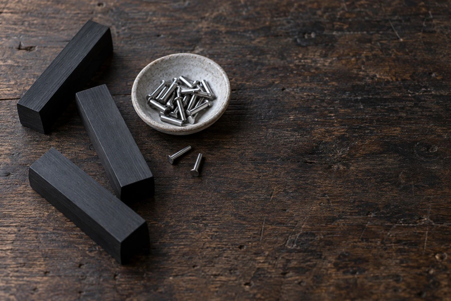 Three plain black handle blanks and a small dish of steel rivet pins on a dark oak workbench