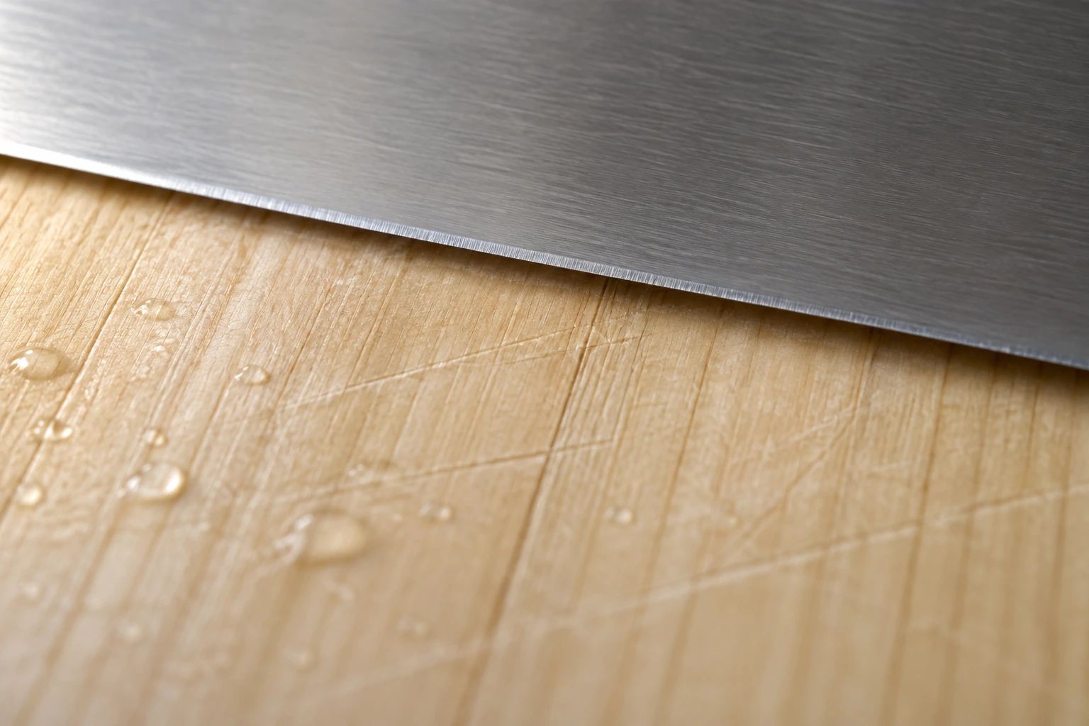 Close-up of a plain steel edge resting lightly on a hinoki board with faint knife marks and water drops