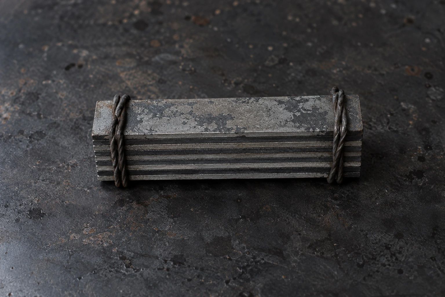 A cold stack of thin steel plates in two tones, bound with wire, on a black iron plate