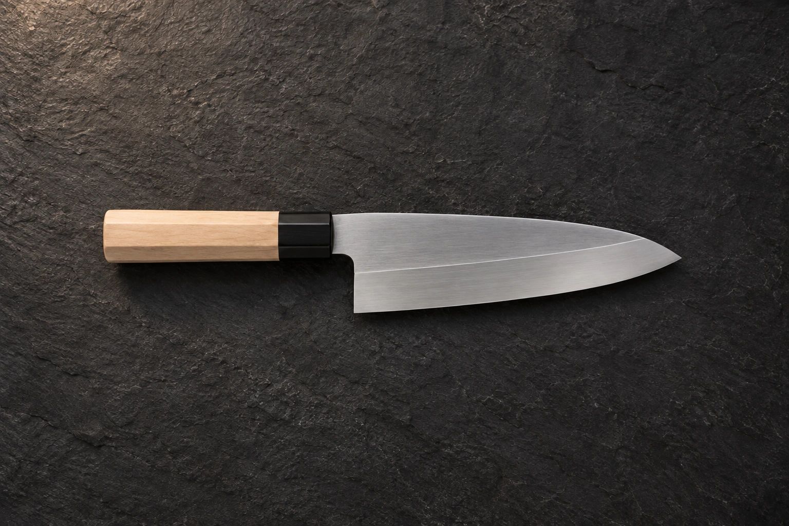 A single-bevel deba knife with a wooden handle on dark slate, seen from above