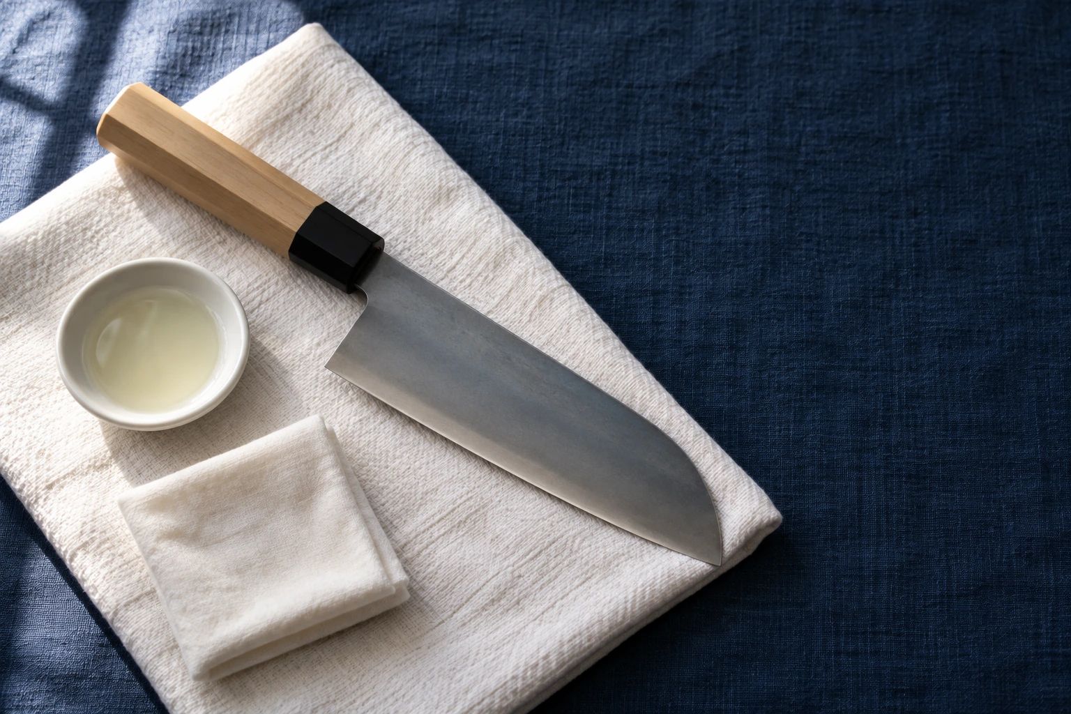 A dried carbon steel santoku on a white towel over indigo cloth, with a dish of camellia oil and a soft cloth