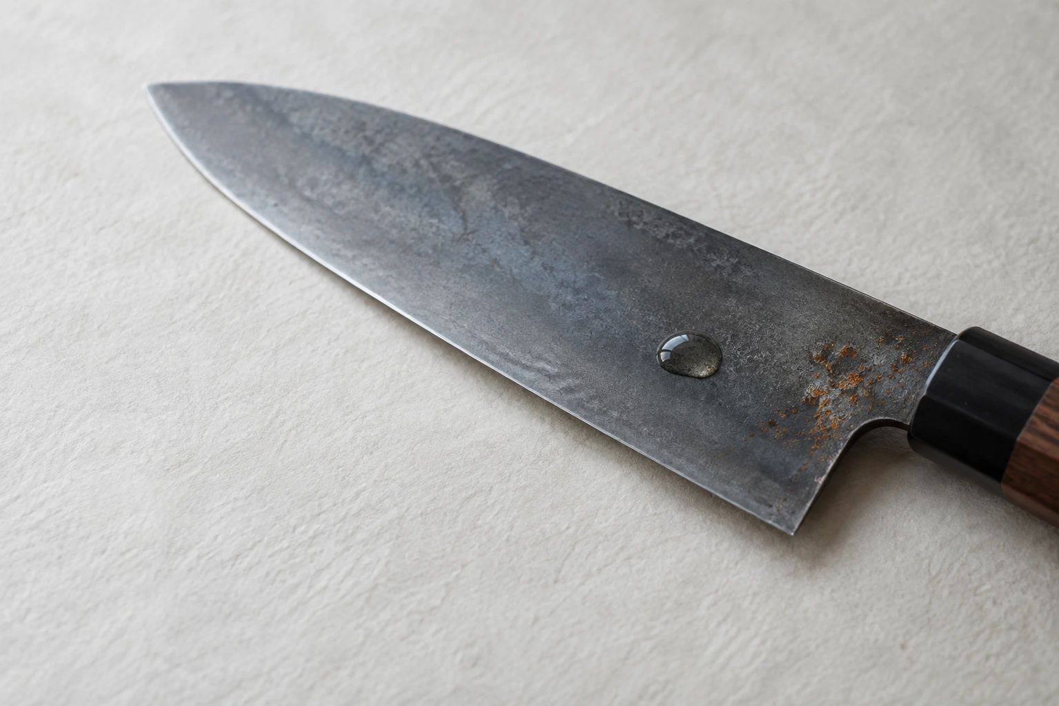 The flat of a carbon steel blade with grey patina, a patch of small orange rust spots and a drop of camellia oil