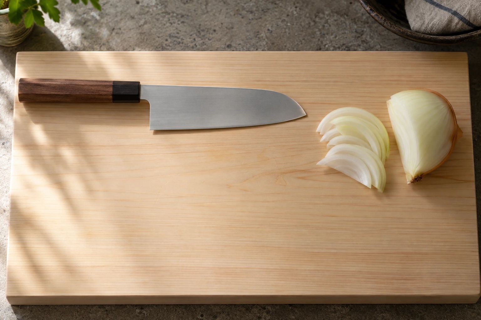 A santoku placed with its handle pointing left on a hinoki board, with a halved and sliced onion on the right