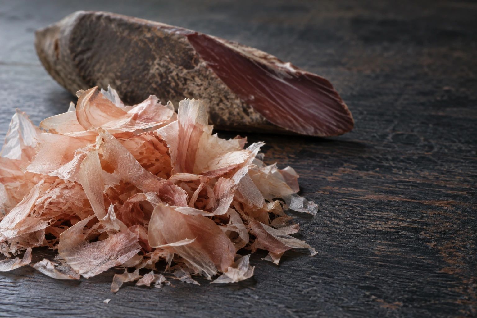 Thin curls of shaved bonito flakes in front of a whole block of dried katsuobushi on dark cedar