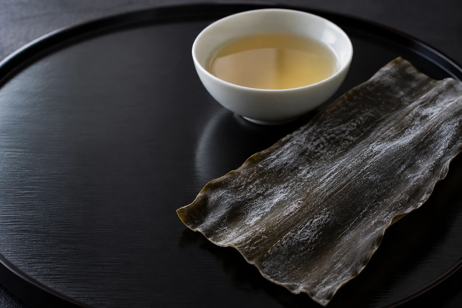 A sheet of dried kombu with a light white bloom and a small bowl of clear dashi on a black lacquer tray