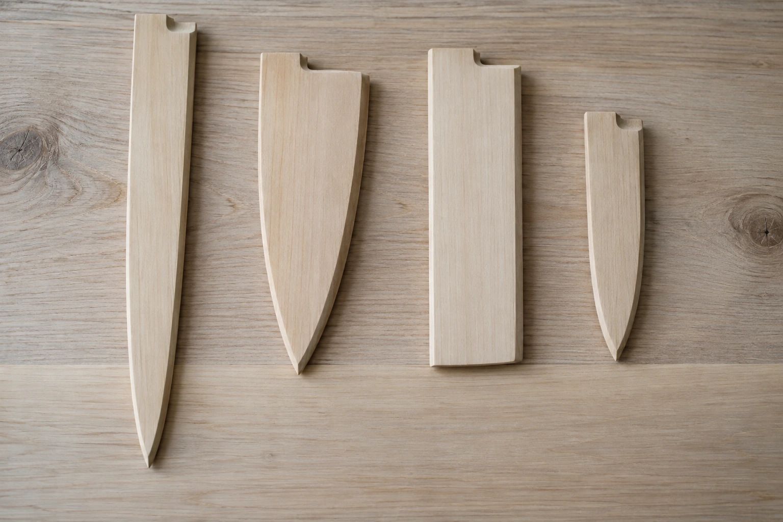 Four empty wooden sayas shaped for a yanagiba, a deba, a nakiri and a petty knife on a pale ash board