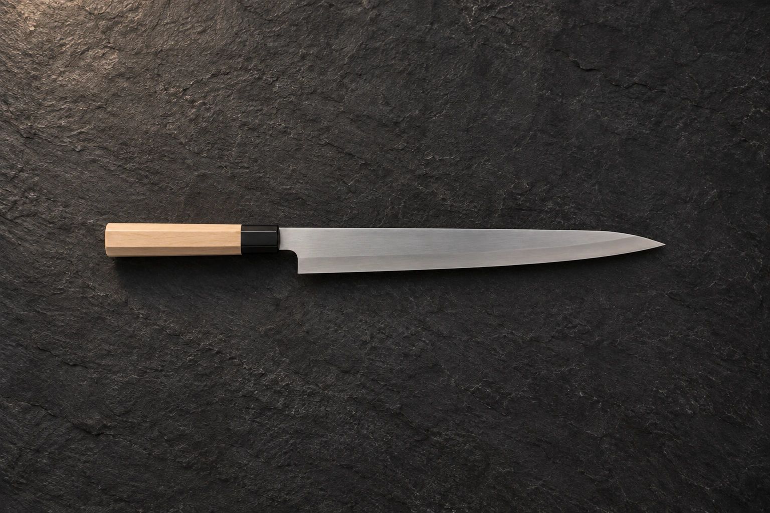A long single-bevel yanagiba with a wooden handle on dark slate, seen from above